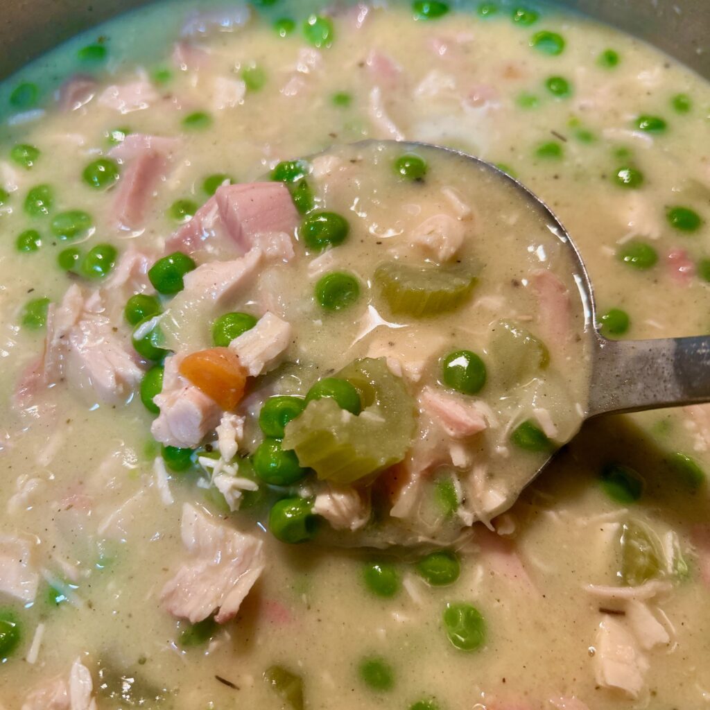 up close picture of simple creamy chicken soup with chicken, green peas, carrots, celery in a pot with a ladle full of soup