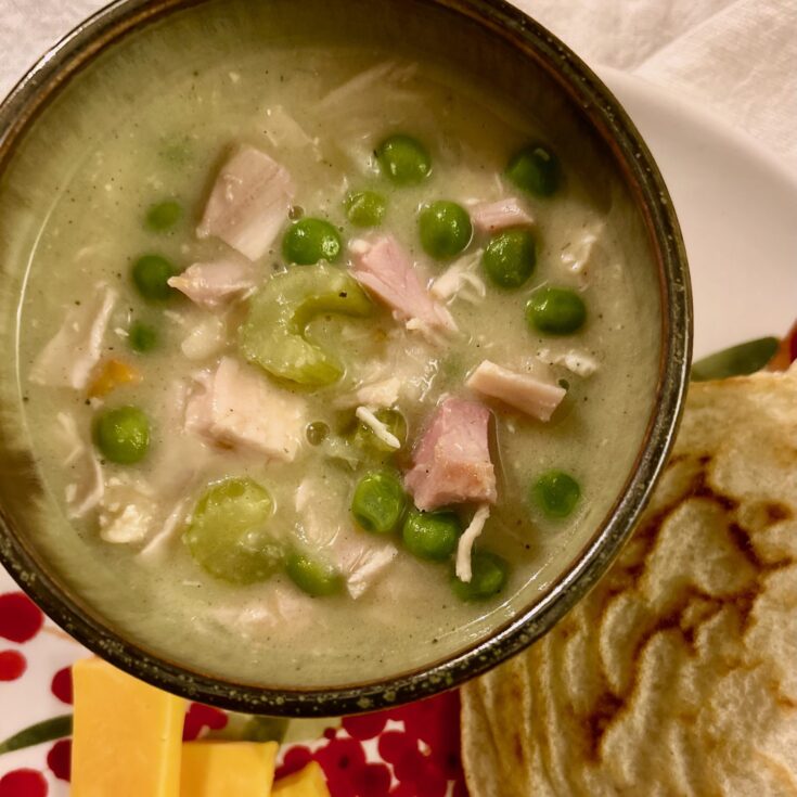 up close picture of simple creamy chicken soup with green peas, celery, carrots, chicken in a pottery bowl on a plate with cheese and flatbread