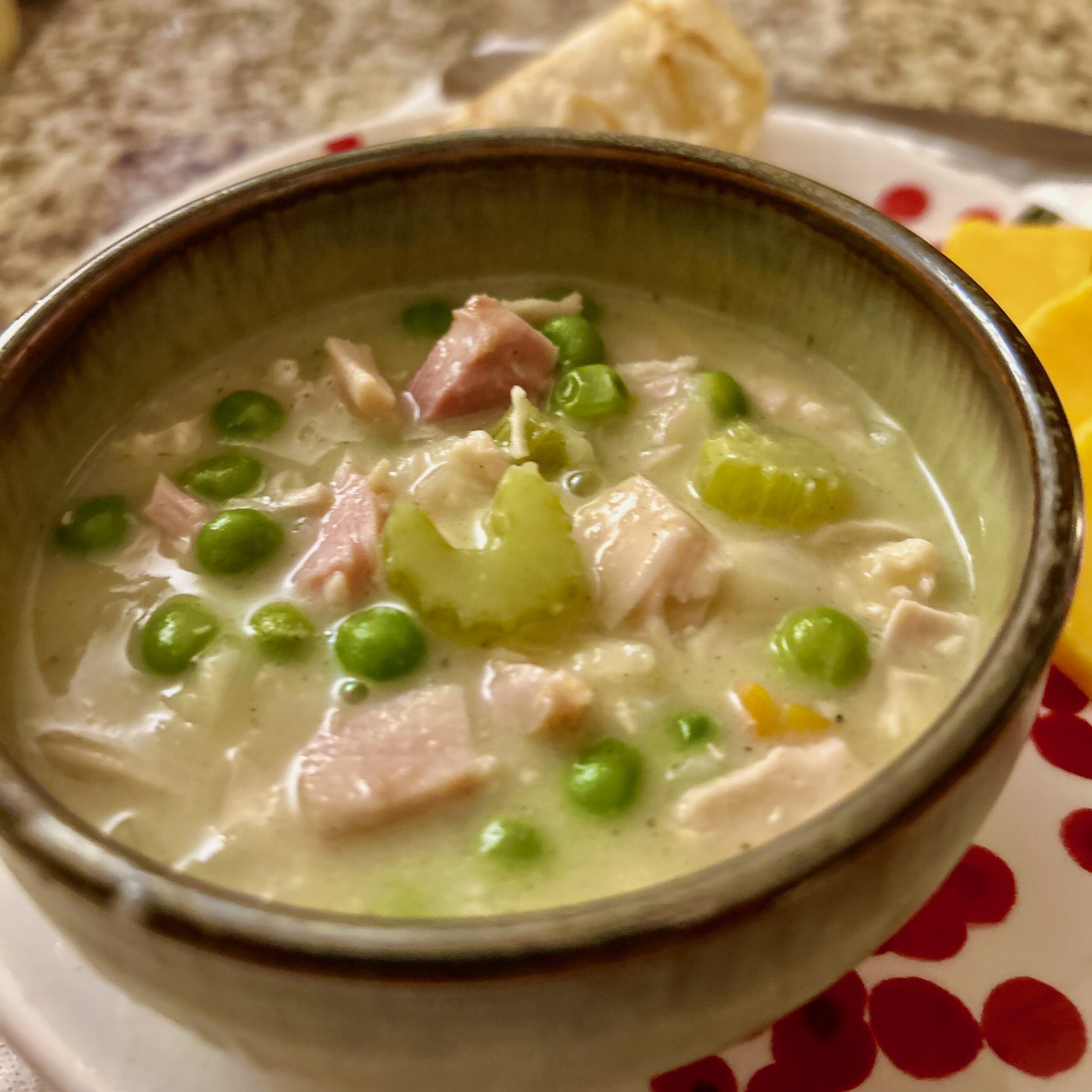close up image of creamy chicken soup with celery, chicken, carrots, and green peas in a pottery bowl on a decorative plate